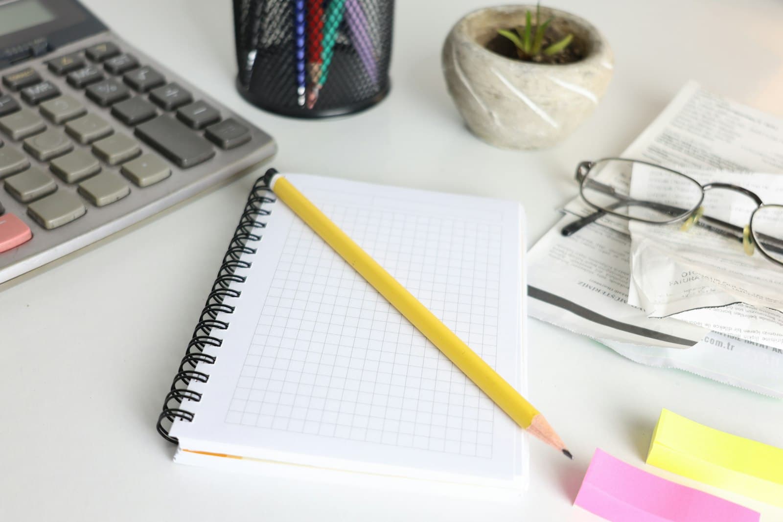 A desk with a calculator, notebook, pencil, glasses, and papers