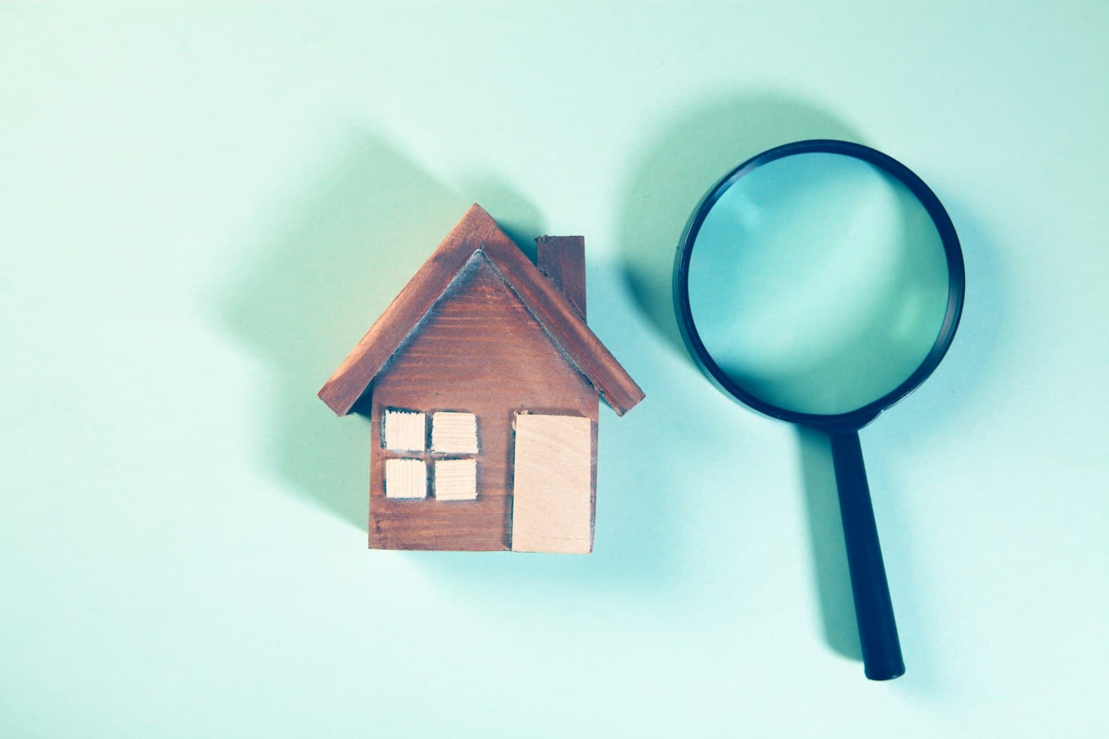 A magnifying glass beside a small wooden house model on a light teal background